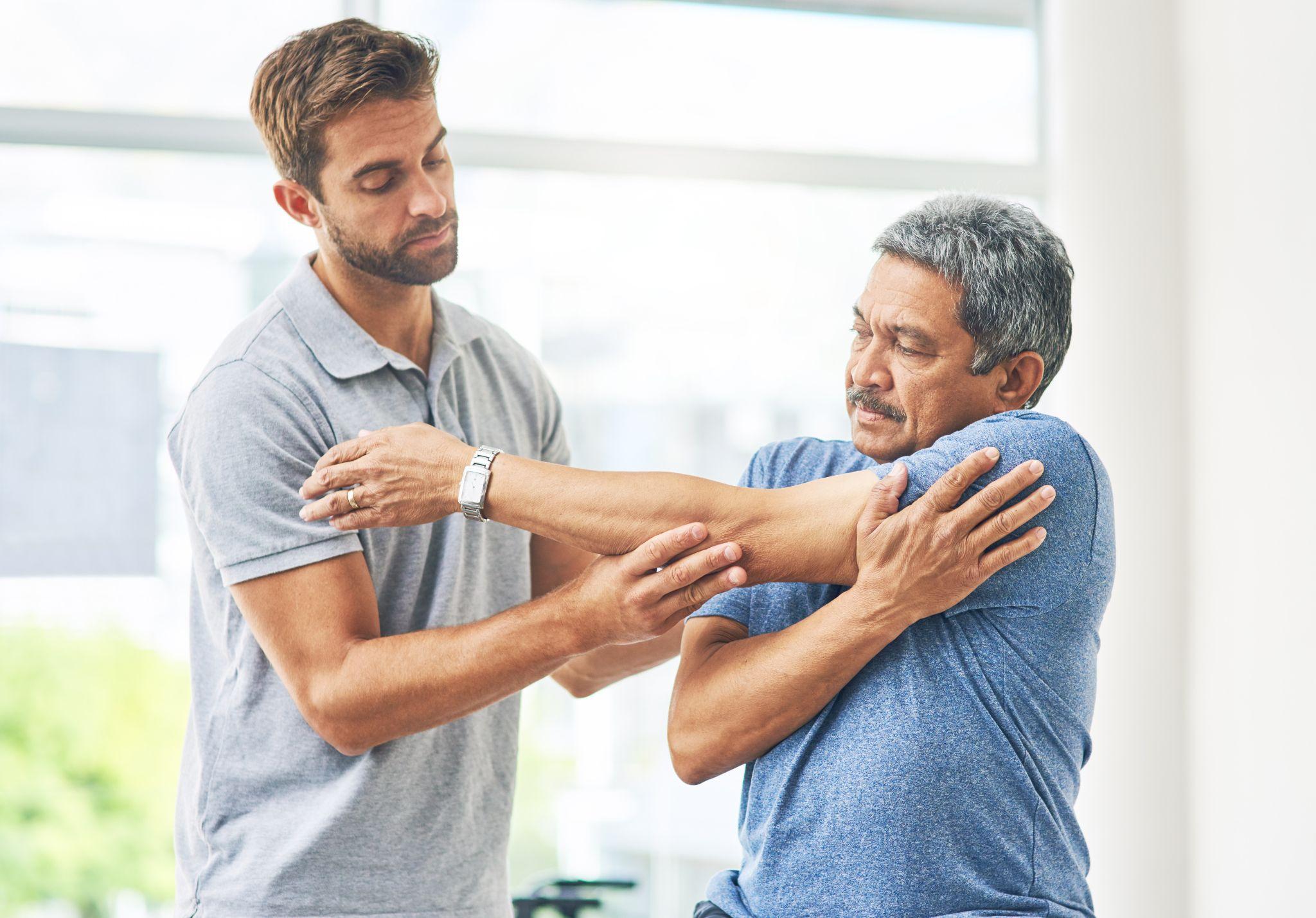 A physiotherapist assisting a senior man with shoulder stretching exercises in a clinical setting.