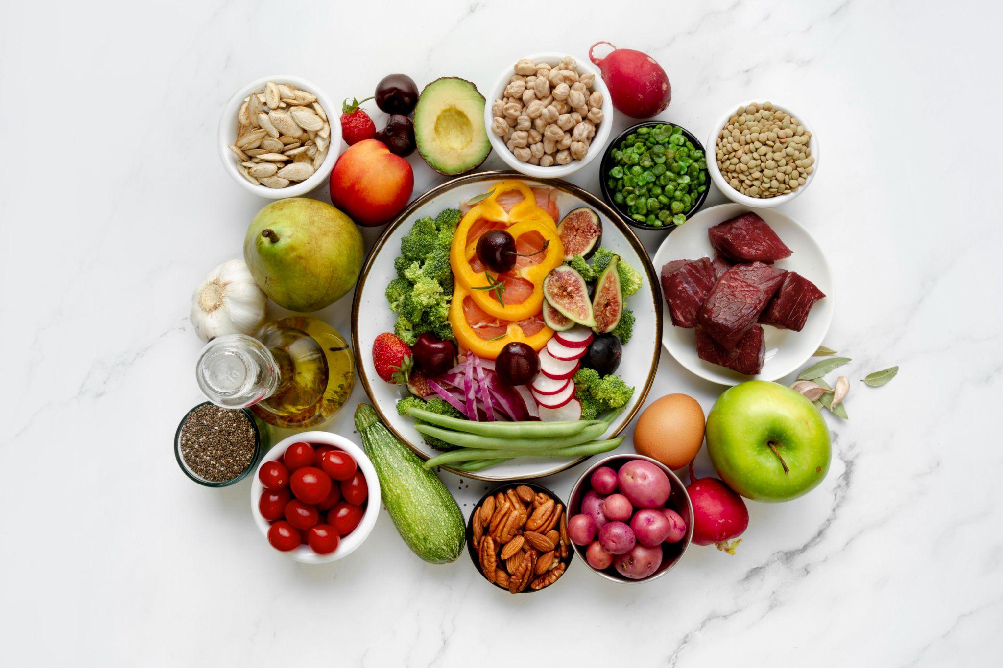 A vibrant top-view image showing a plate filled with colorful vegetables like broccoli, bell peppers, tomatoes, figs, and radishes, surrounded by bowls of nuts, beans, lentils, avocado, fruits and cherry tomatoes. The layout represents a healthy, nutrient-rich, whole-foods diet.