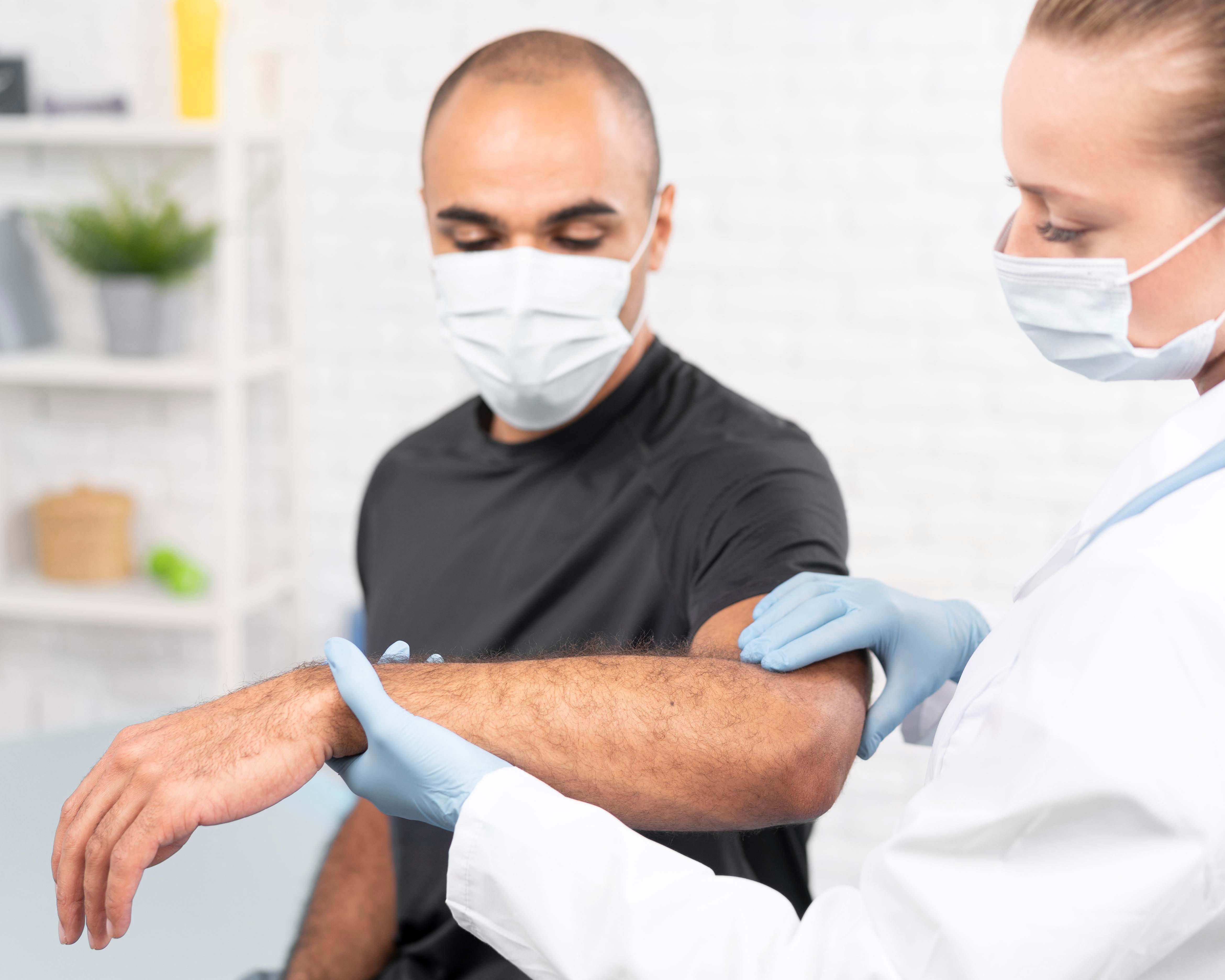 Female physiotherapist examining a man's elbow while both wear medical masks.