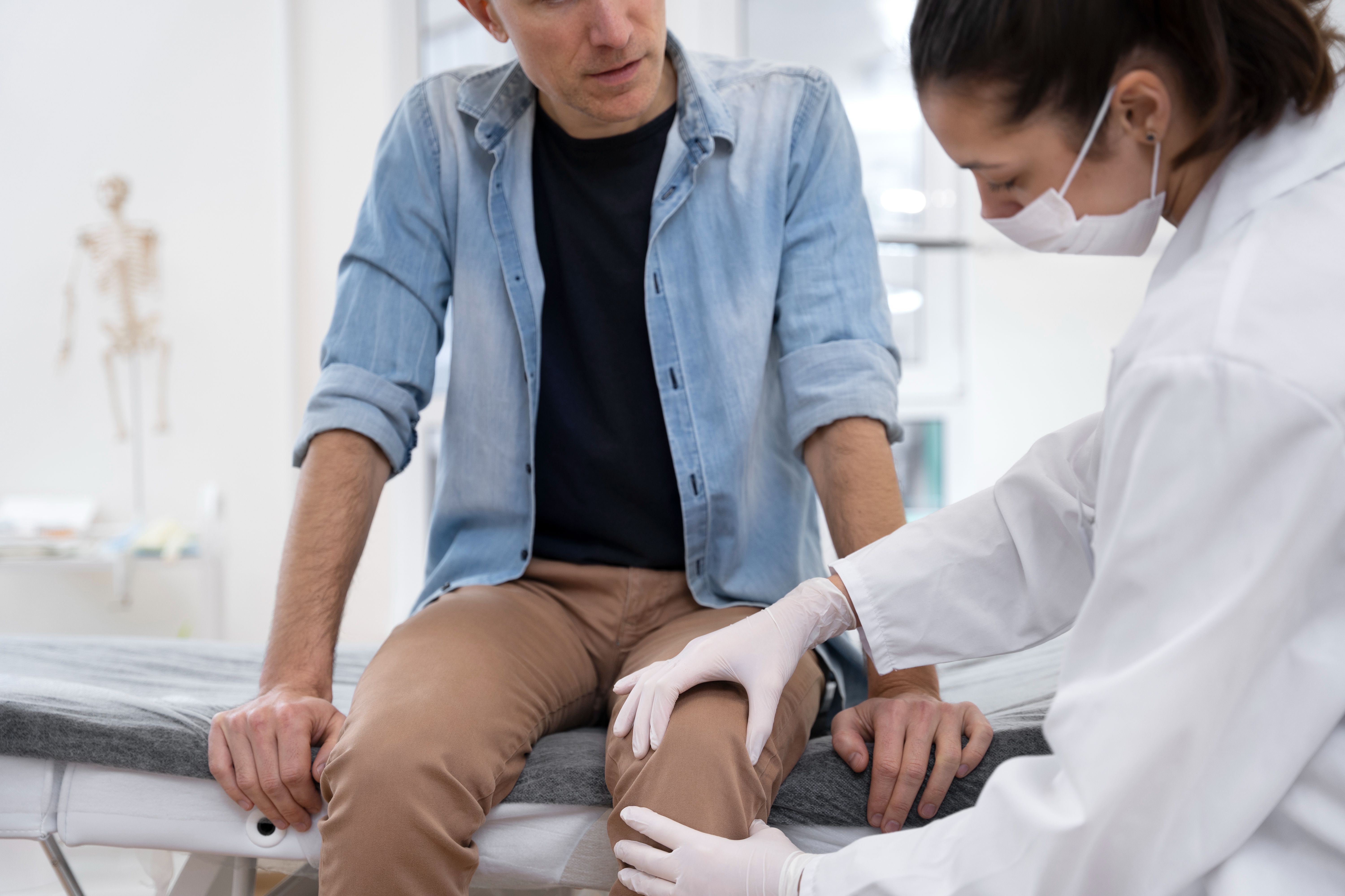 A male patient sits on an examination table while a female doctor, wearing a mask and gloves, examines his knee in a medical clinic.