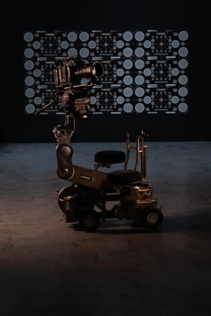 A cinema camera on a dolly arm in front of a wall-sized Hawk lens test chart in a dark test room
