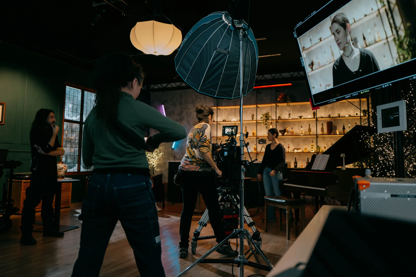 A film crew shooting a scene on the dressed bar set with a grand piano and backlit shelves inside Hawk London's space in Stratford, with the shot on a monitor