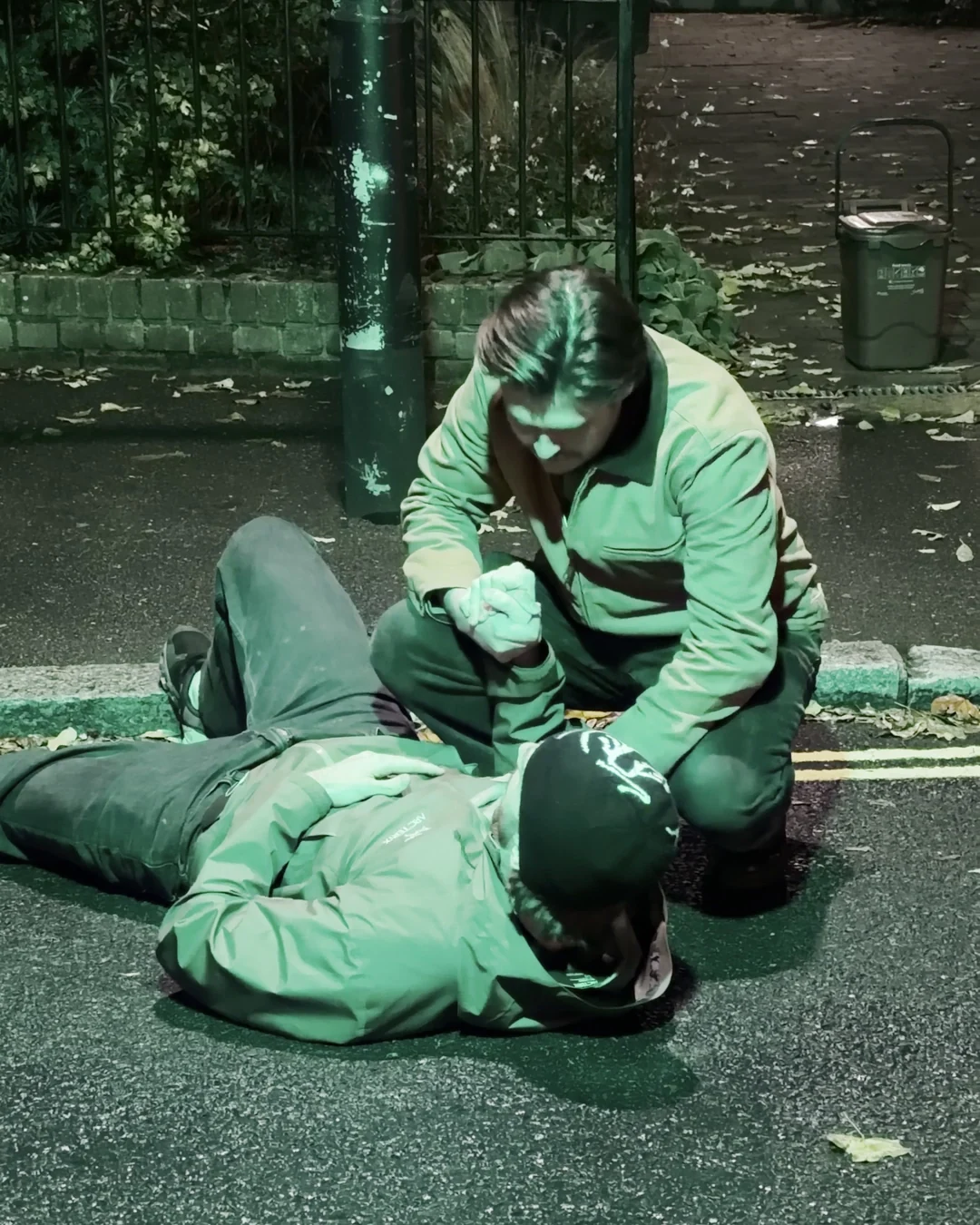 Actor kneeling over a man lying in the road, giving mock CPR beside a parked Lime bike at night