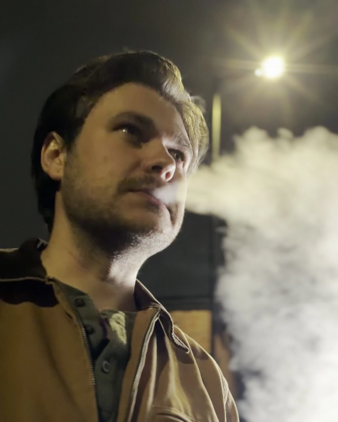 Actor in profile exhaling smoke under a single warm street light at night, the background falling into darkness