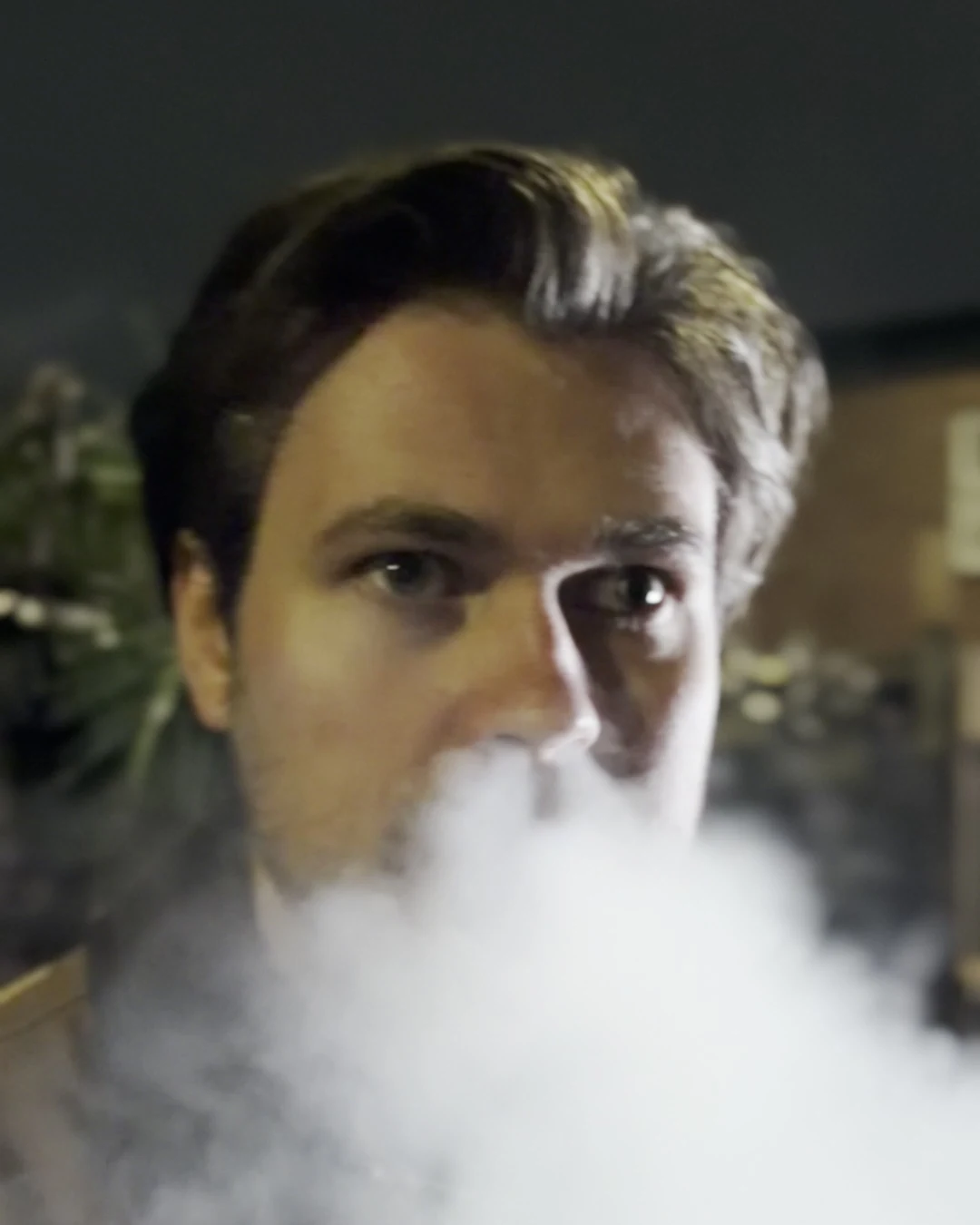 Close-up of an actor breathing a thick cloud of smoke towards the lens under a warm street light