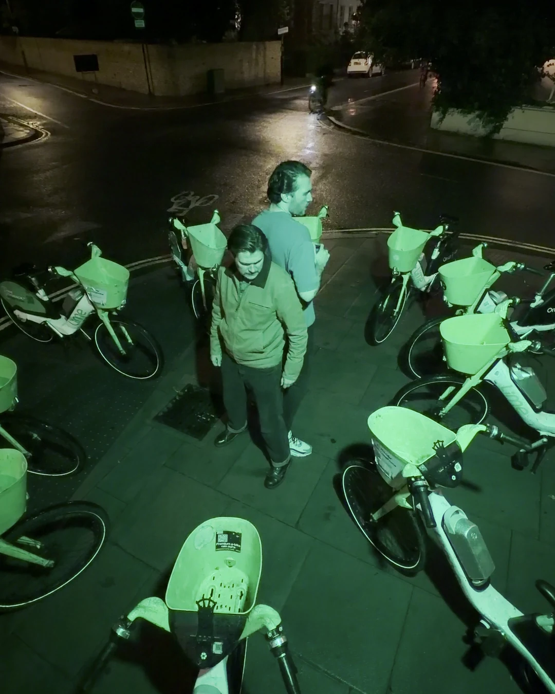 High angle of two actors standing among dozens of parked Lime bikes washed in green light at night