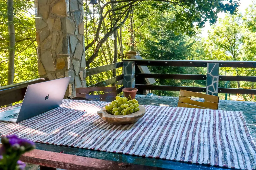 Covered stone terrace with a wooden table holding a laptop and a bowl of grapes, surrounded by trees