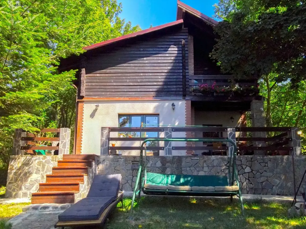 Two-storey chalet with dark wood cladding above a stone-clad ground floor, seen from the garden with a porch swing and a sun lounger on the lawn