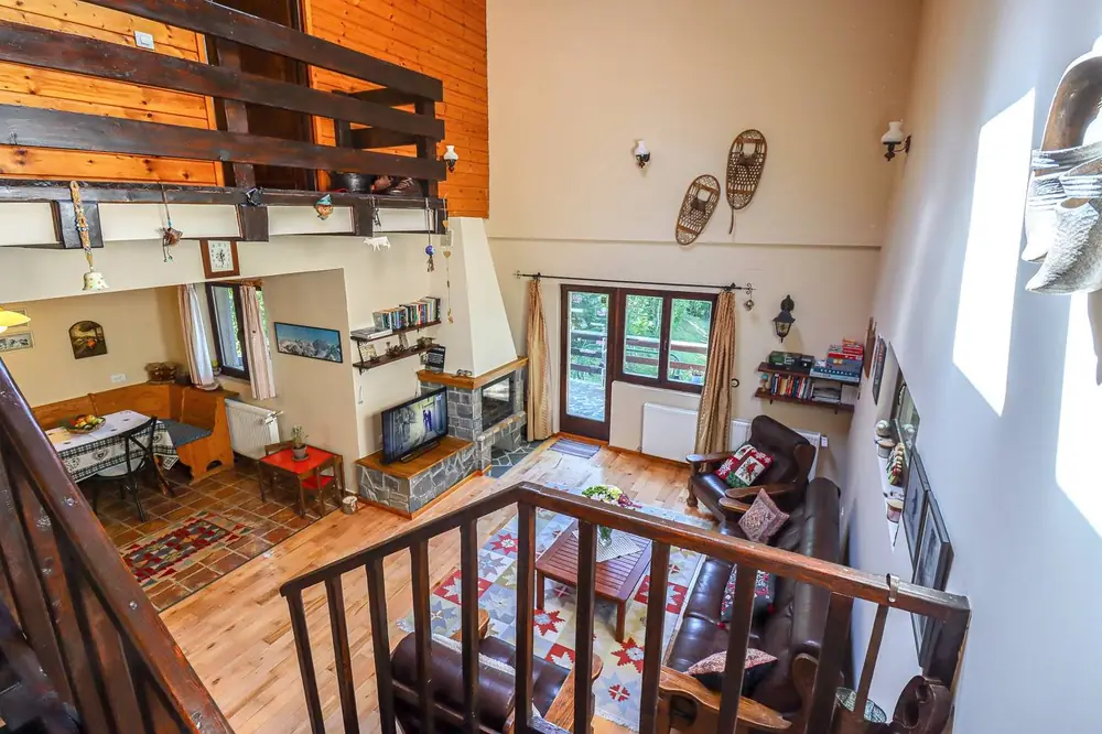 Overlook from the wooden loft railing down into the double-height living room, with leather sofas, a patterned rug, and exposed timber beams