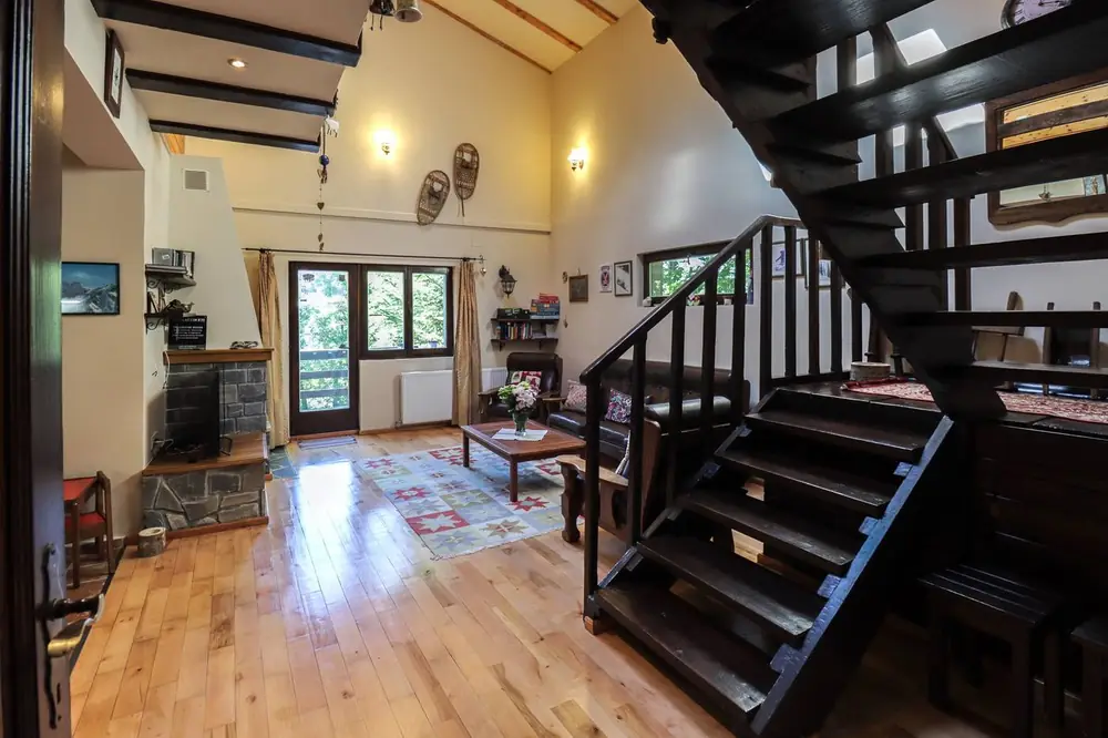 Living room with a dark wooden staircase, exposed beam ceiling, a corner wood-burning stove, and glass doors opening onto the porch