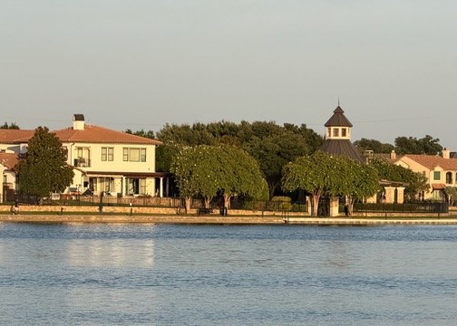 Golden-hour lakeside homes reflected in calm water with mature trees and a walking path