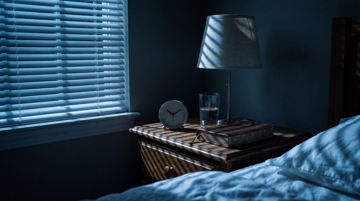 A bedside table at night with an analogue clock, a glass of water and a closed Bible, lit by cold moonlight through blinds