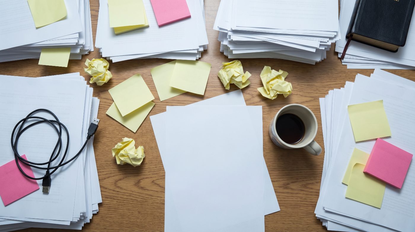 A cluttered desk covered in papers and sticky notes, a Bible half-buried in the mess
