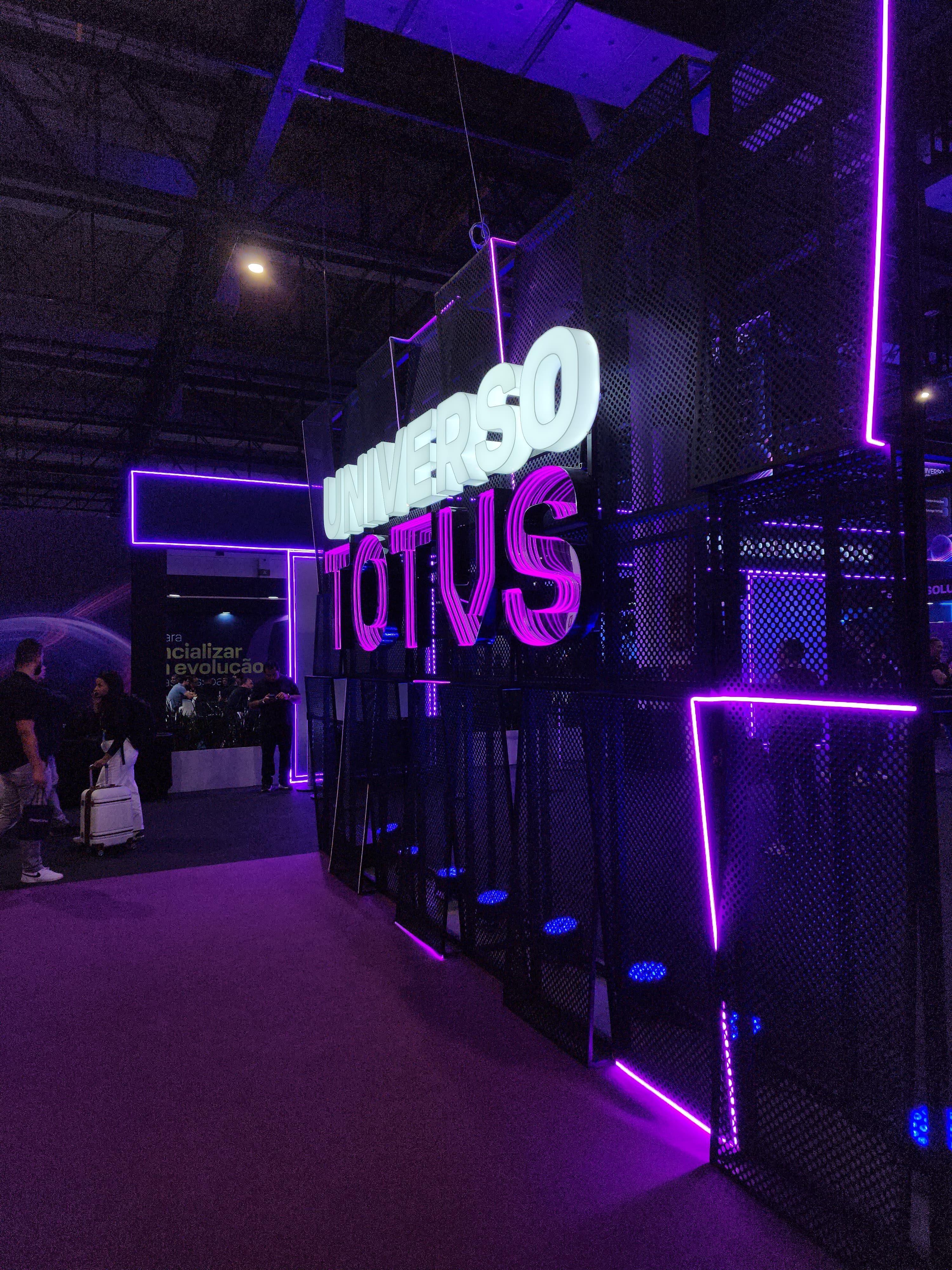 Estande da TOTVS com letreiro iluminado "Universo TOTVS" em neon roxo e branco durante evento de tecnologia e negócios.