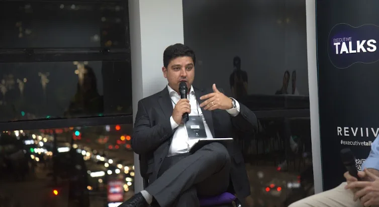 Renato Mattos mediando painel no Executive Talks, com microfone em mãos e bloco de anotações, diante de uma janela com vista noturna da cidade.