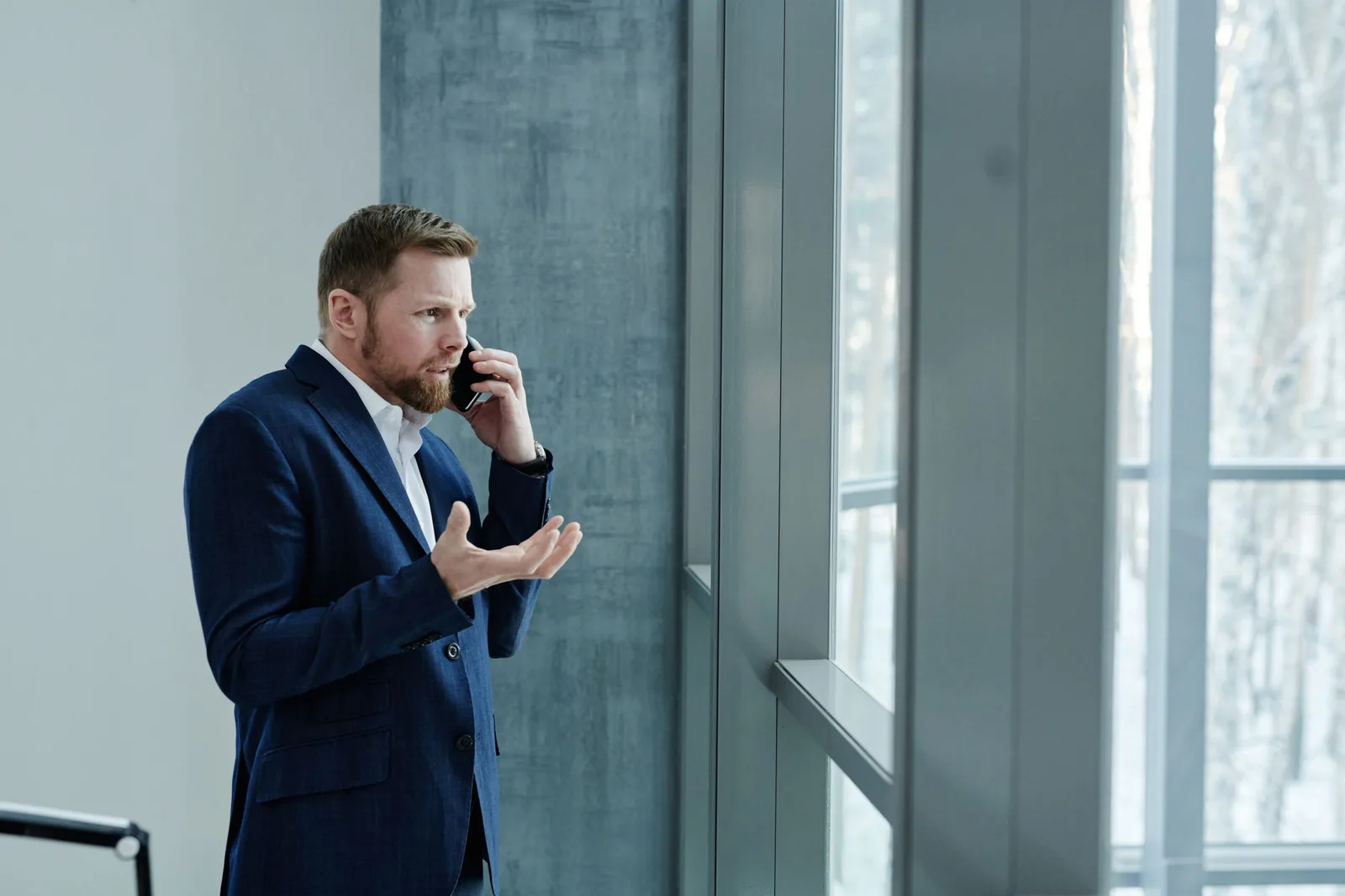 Homem de terno azul gesticulando preocupado ao telefone perto de uma janela grande, ilustrando a reação de uma vítima ao receber chamadas de golpes virtuais por clonagem de voz.