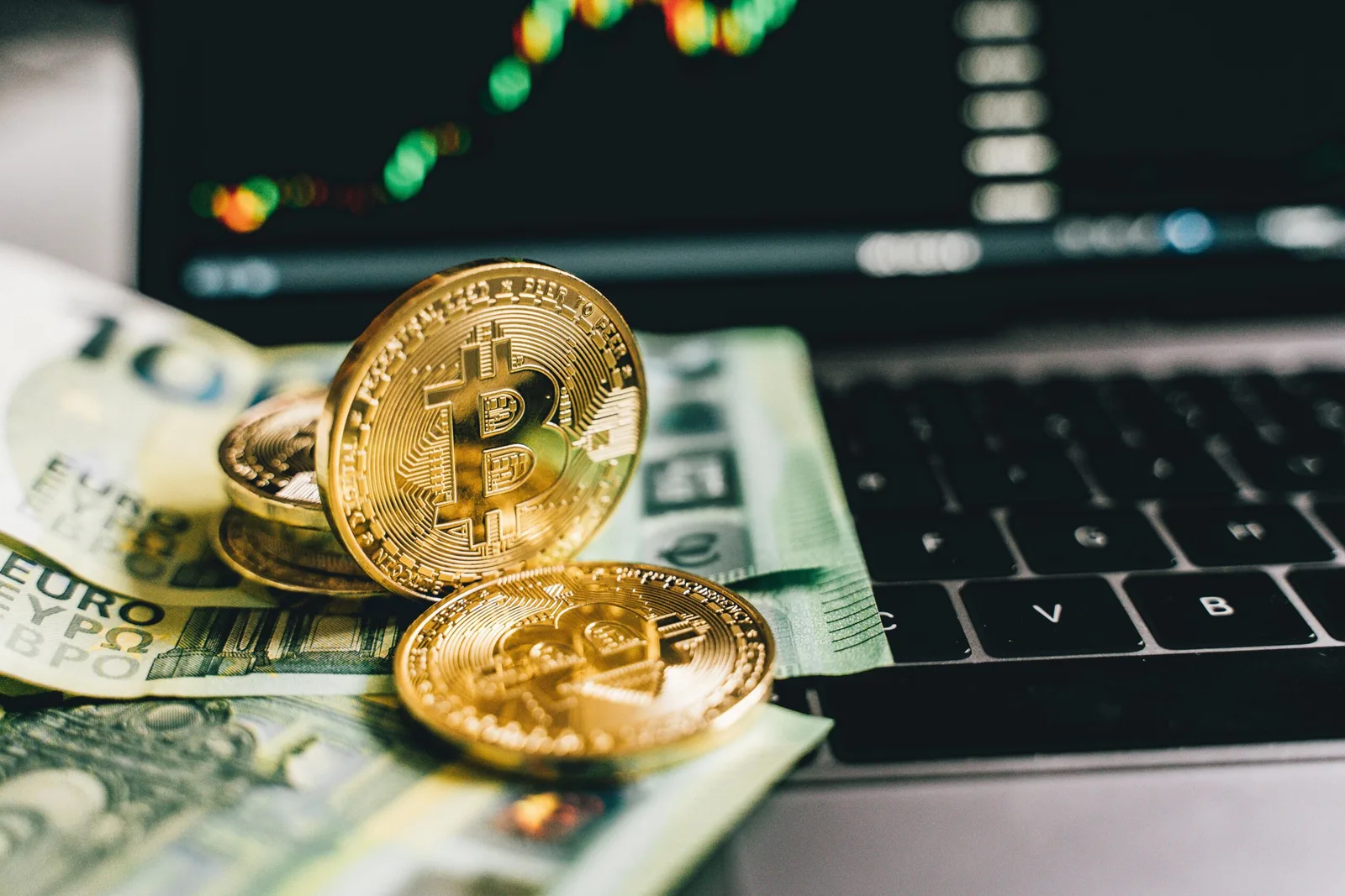 Fotografia em close sobre um notebook cinza, mostrando moedas douradas de Bitcoin (uma em pé e outras deitadas) apoiadas sobre um monte de cédulas de dólar e real. Ao fundo, desfocado, um monitor exibe um gráfico de barras e linha de mercado financeiro.