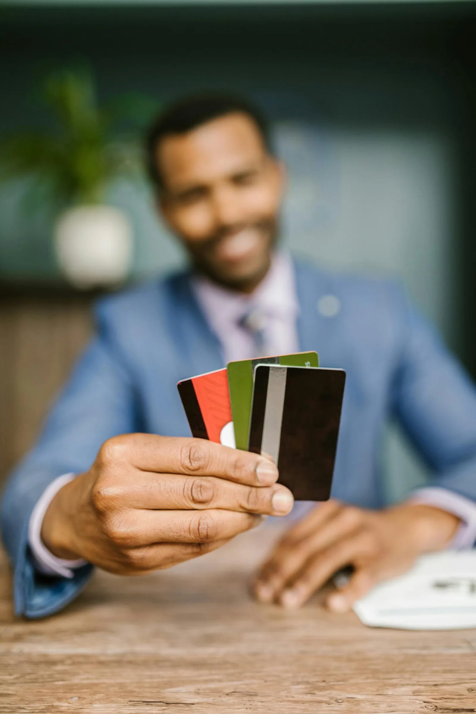 Close-up das mãos de um homem negro segurando quatro cartões de crédito e débito diferentes (preto, verde e vermelho) em direção à câmera. Ele veste um terno azul e camisote de blazer, com um fundo desfocado de escritório e uma planta.