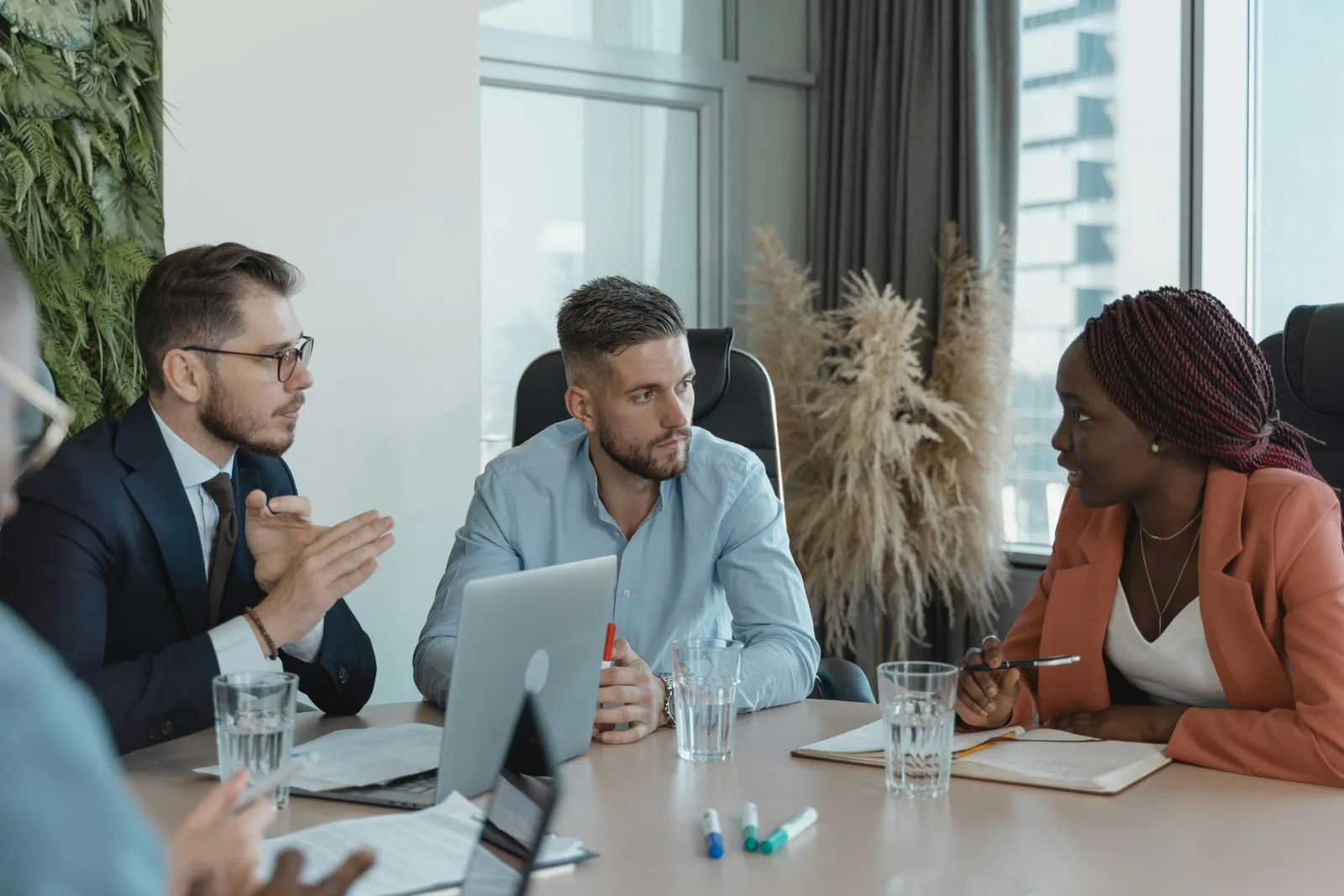 Três executivos — uma mulher negra de blazer laranja e dois homens — em uma reunião estratégica de negócios. Eles estão sentados em uma mesa com laptops, cadernos e copos de água em um escritório moderno, discutindo a implementação de Inteligência Artificial.
