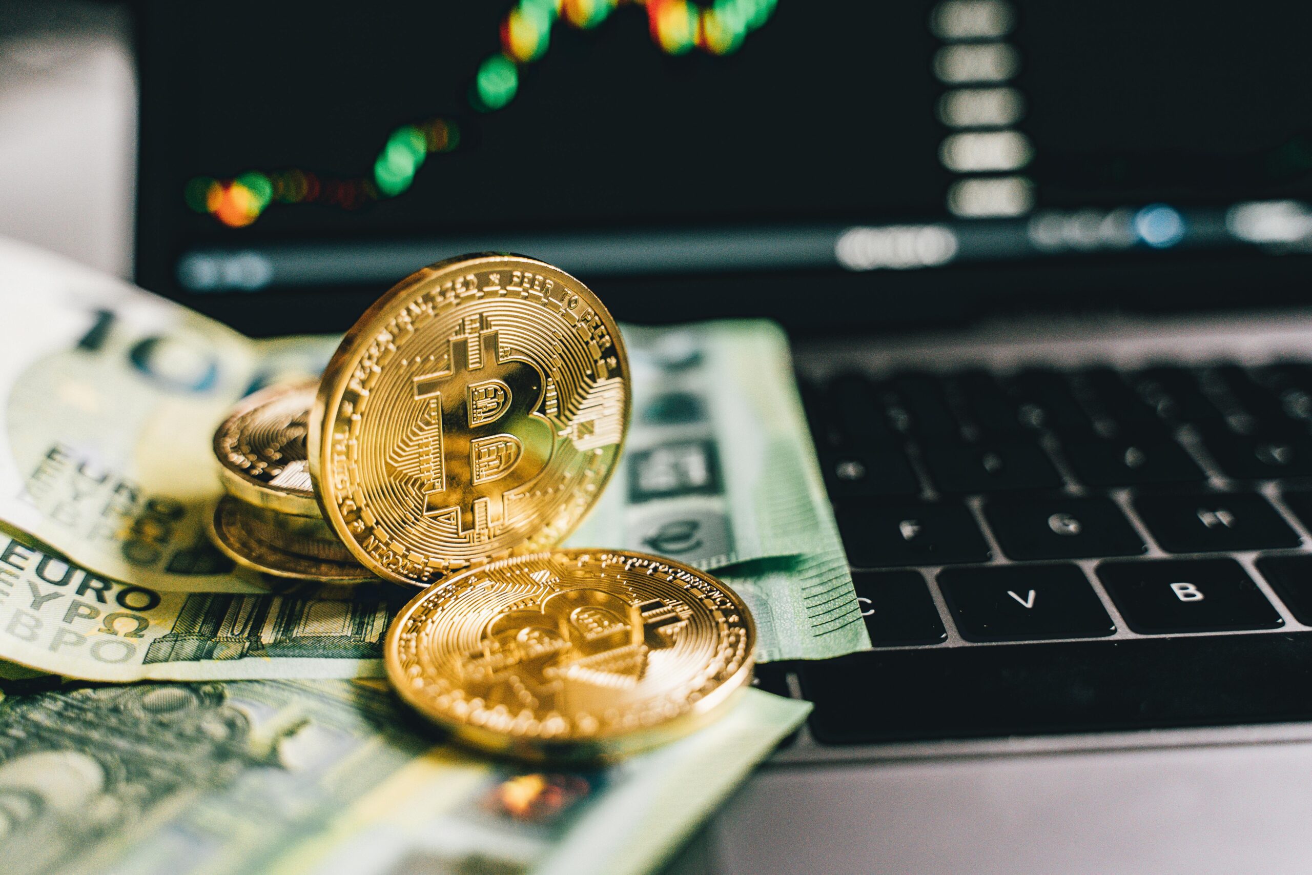 Fotografia em close sobre um notebook cinza, mostrando moedas douradas de Bitcoin (uma em pé e outras deitadas) apoiadas sobre um monte de cédulas de dólar e real. Ao fundo, desfocado, um monitor exibe um gráfico de barras e linha de mercado financeiro.