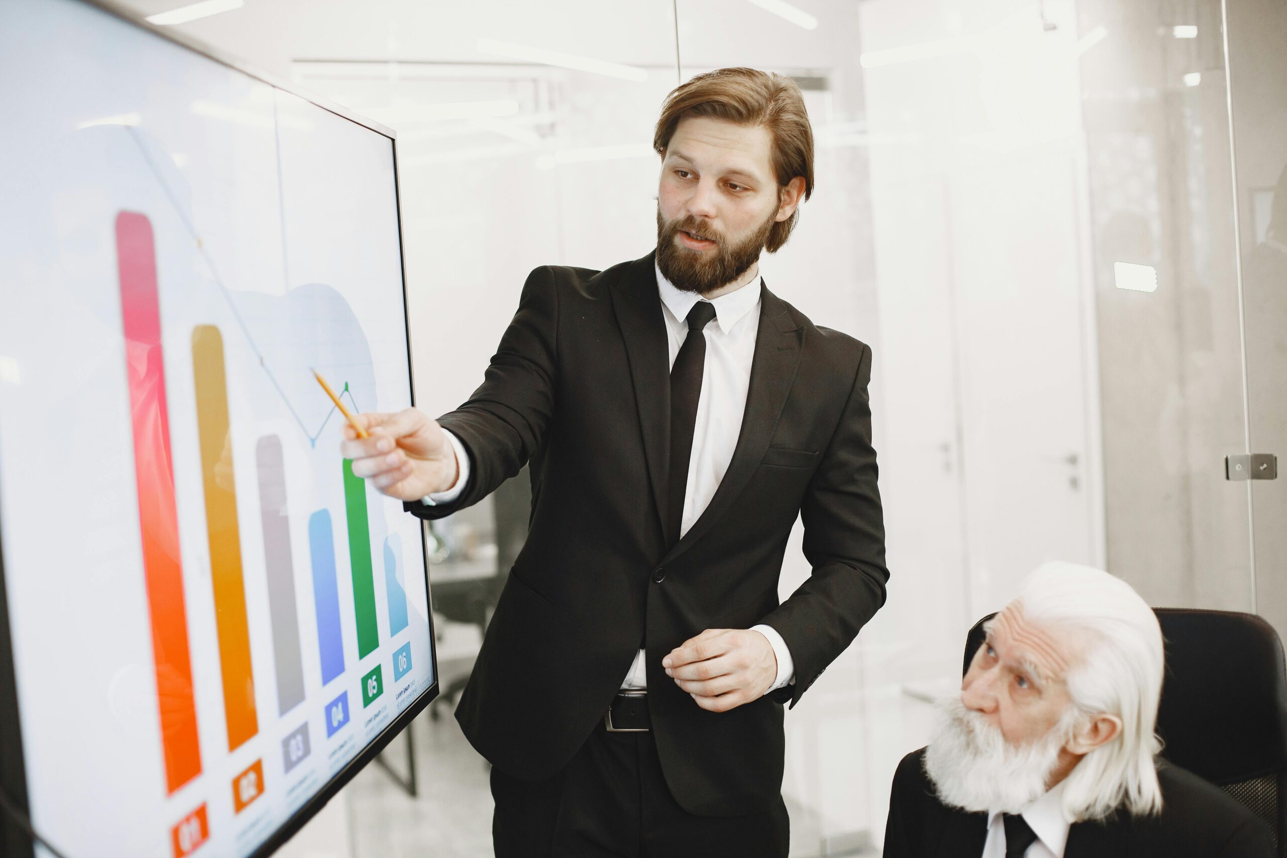 Homem de terno preto apontando com um lápis para um gráfico de barras coloridas em uma tela, apresentando resultados financeiros da Anthropic para um executivo sênior de cabelos e barba branca.