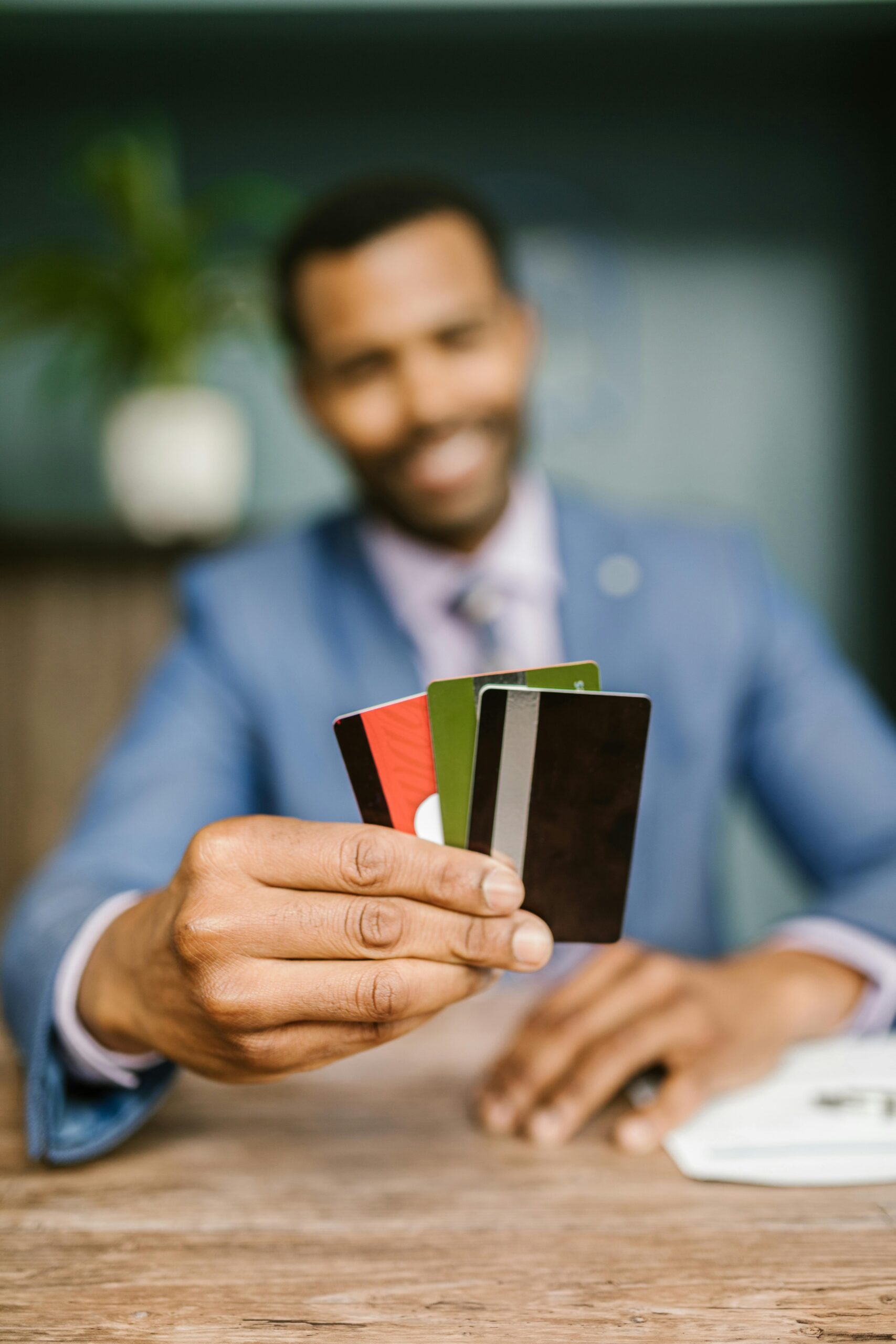 Close-up das mãos de um homem negro segurando quatro cartões de crédito e débito diferentes (preto, verde e vermelho) em direção à câmera. Ele veste um terno azul e camisote de blazer, com um fundo desfocado de escritório e uma planta.