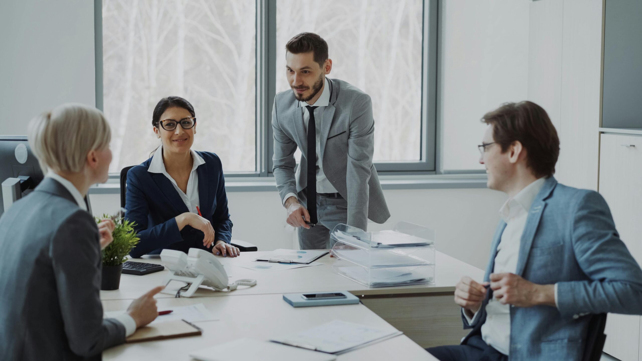 Uma equipe de quatro executivos — duas mulheres e dois homens — reunida em uma sala de conferências moderna. Um homem de terno cinza está em pé gesticulando para documentos, enquanto os outros estão sentados ao redor de uma mesa colaborando. A cena representa a governança humana necessária para aplicar inteligência de negócios.