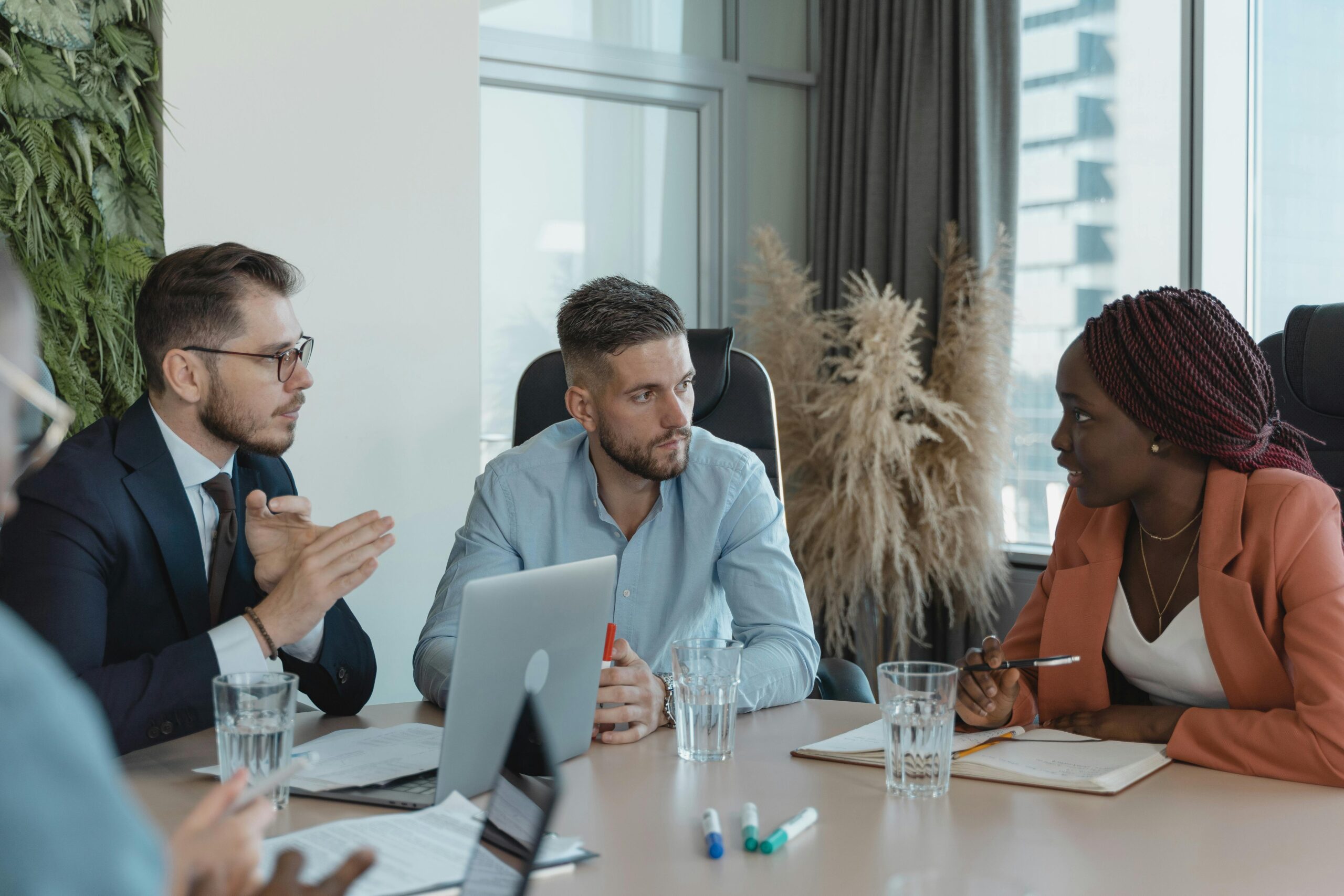 Três executivos — uma mulher negra de blazer laranja e dois homens — em uma reunião estratégica de negócios. Eles estão sentados em uma mesa com laptops, cadernos e copos de água em um escritório moderno, discutindo a implementação de Inteligência Artificial.