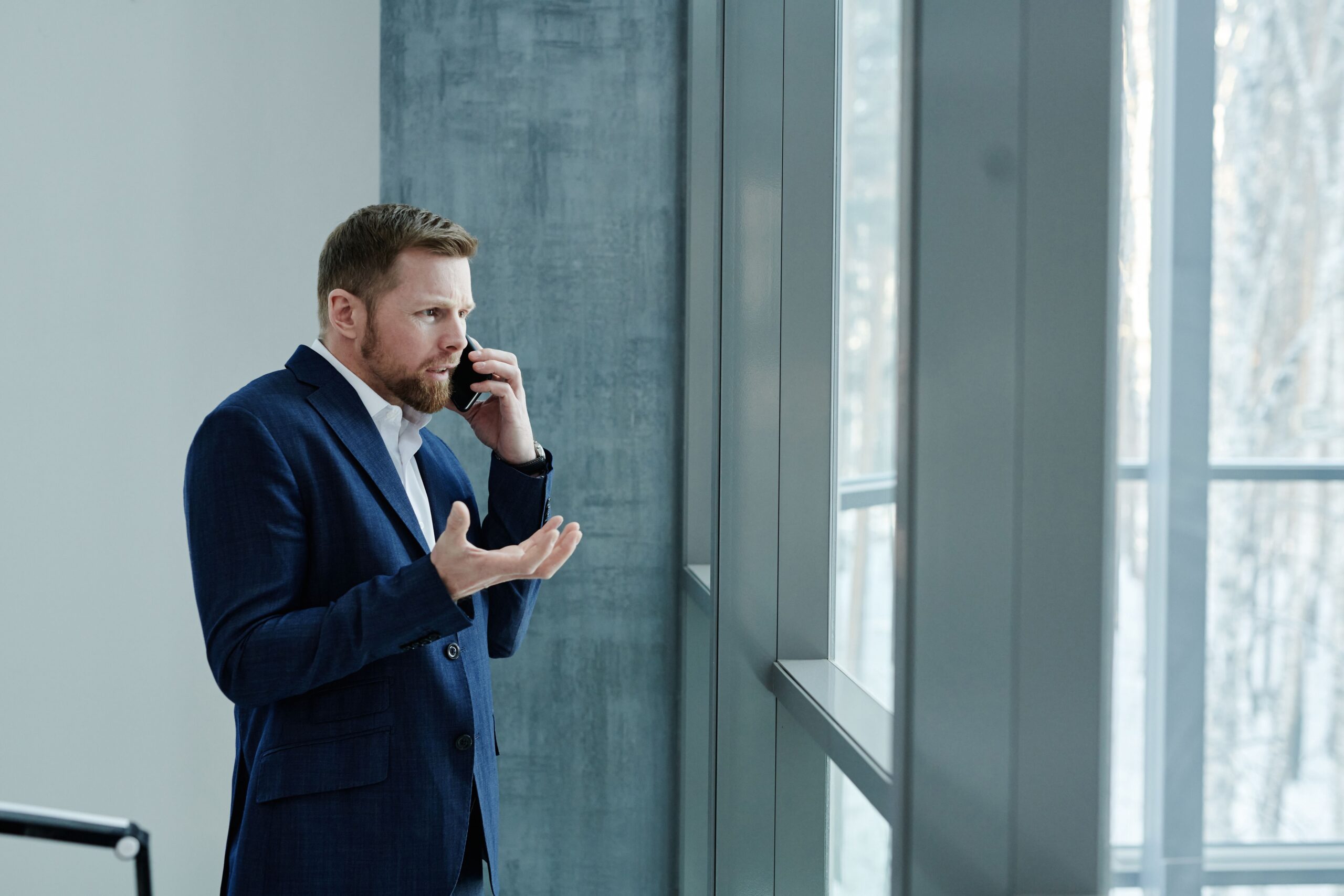 Homem de terno azul gesticulando preocupado ao telefone perto de uma janela grande, ilustrando a reação de uma vítima ao receber chamadas de golpes virtuais por clonagem de voz.