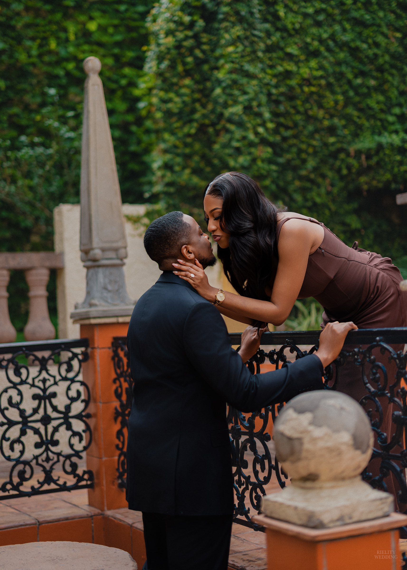 Woman in a brown satin dress leaning over an ornate iron railing to kiss her partner in a garden