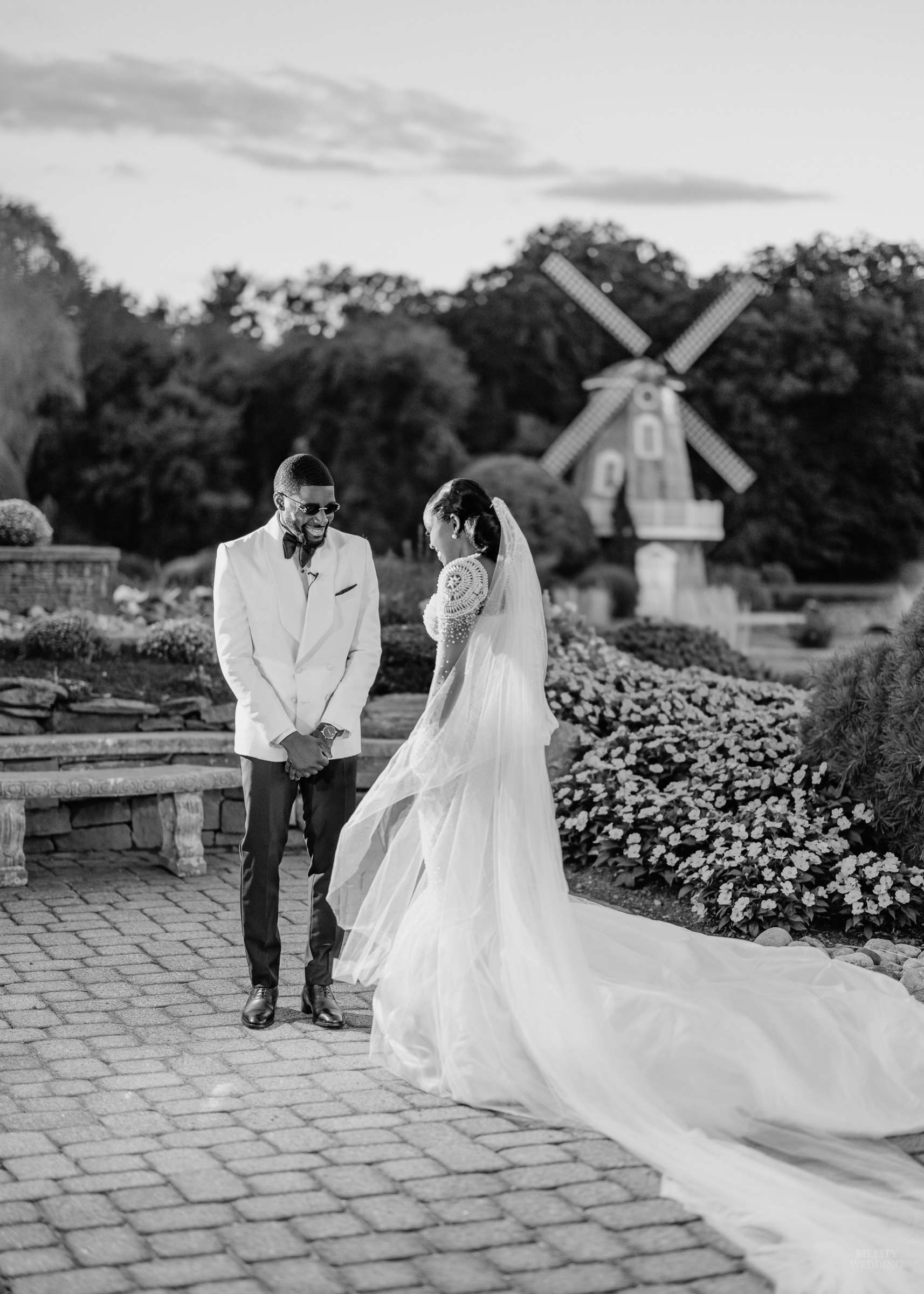 Black and white photo of a bride in a long cathedral veil and a groom in a white tuxedo jacket on a garden path with a windmill behind them