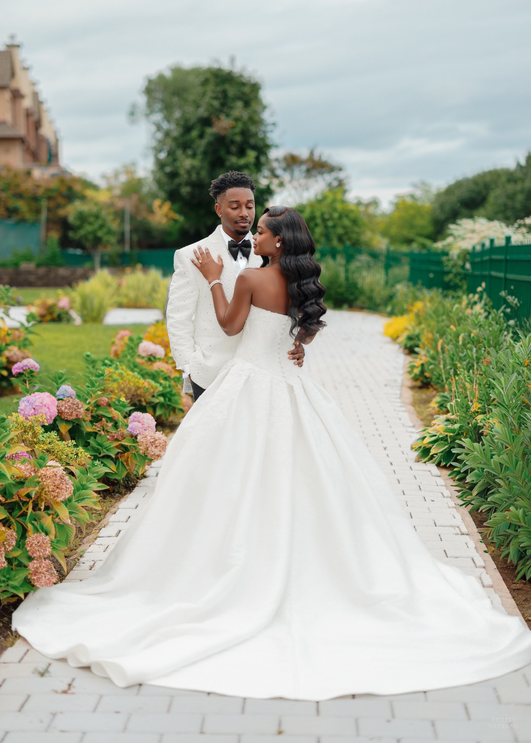 Bride in a strapless ball gown embracing her groom in a white tuxedo jacket on a garden path lined with hydrangeas