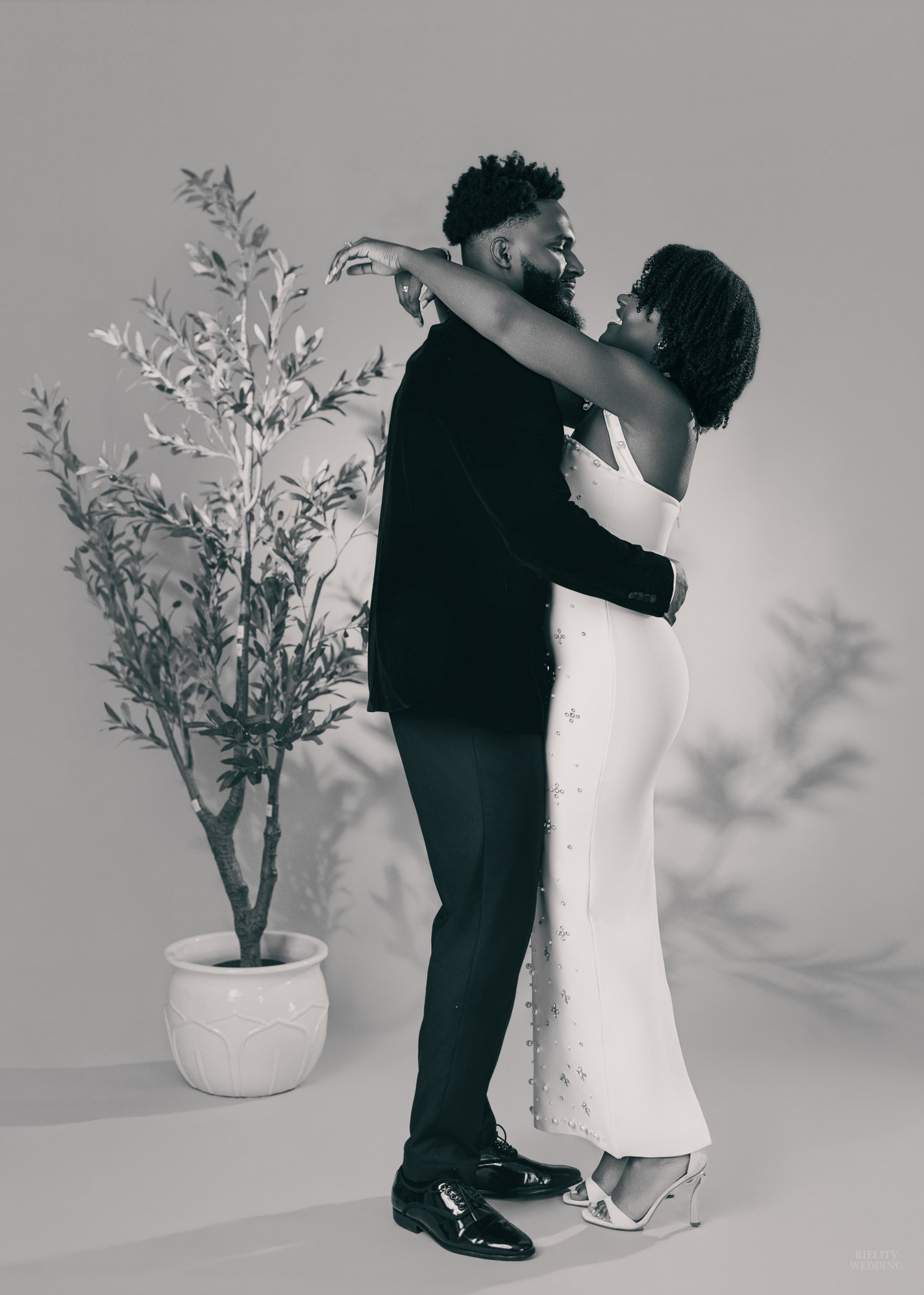Black and white studio portrait of a couple embracing beside a potted olive tree