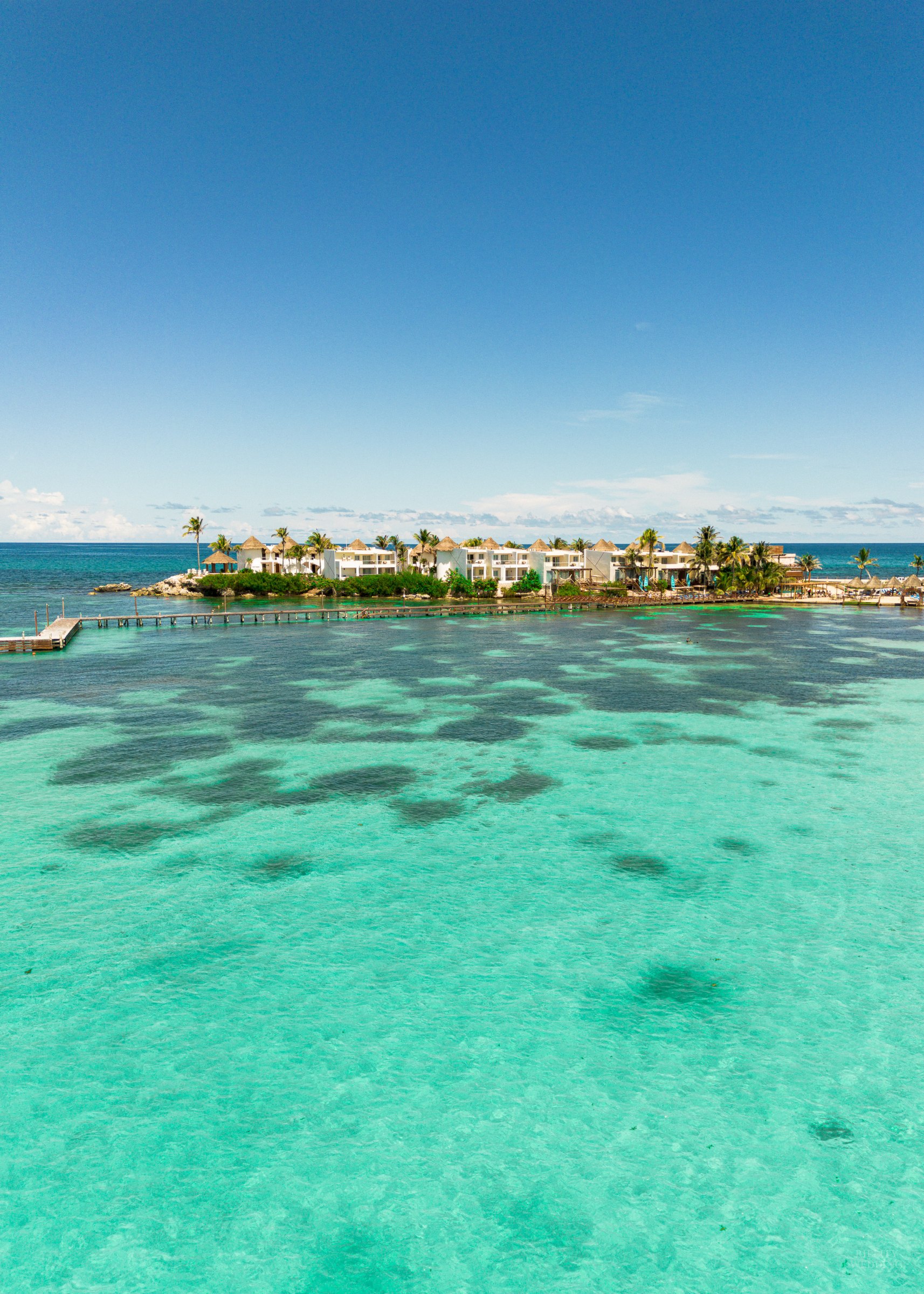 Aerial view of turquoise ocean water and a palm-lined island resort under a clear blue sky