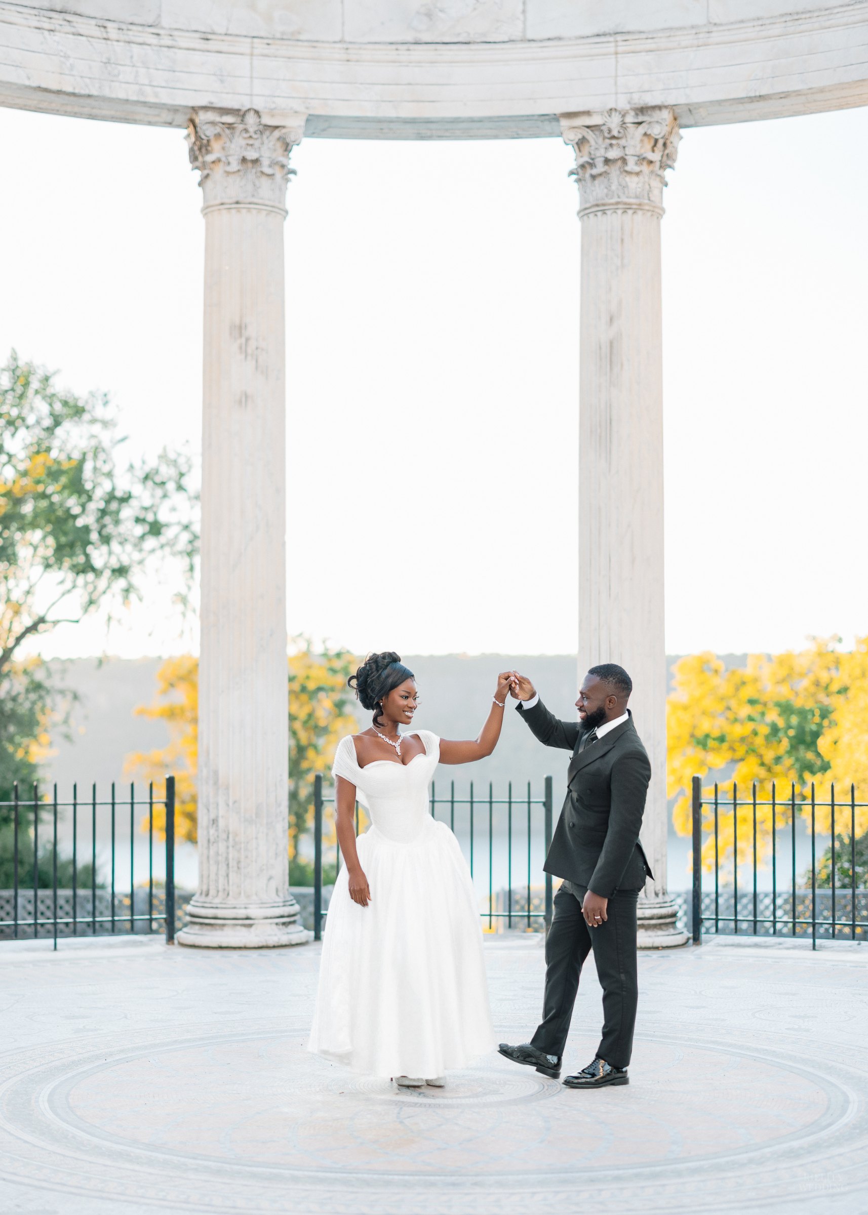 Groom twirling his bride in a tea-length white dress beneath a white marble column rotunda with autumn trees