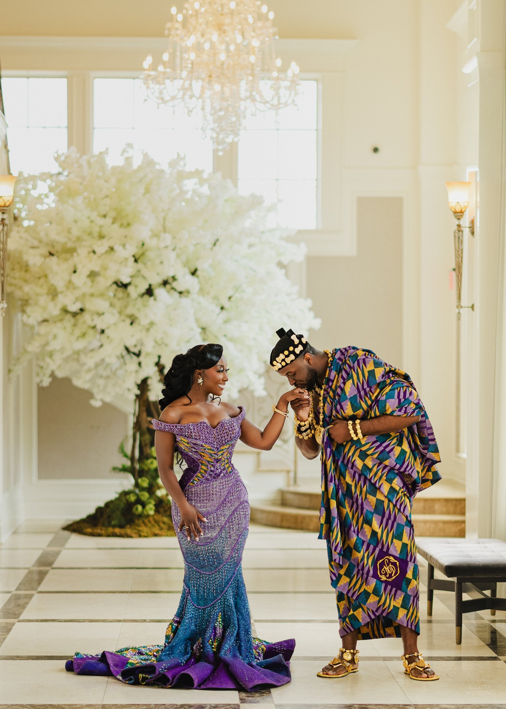 Groom in kente cloth kissing the hand of his bride in a purple and blue gown in a grand hall with a white floral tree