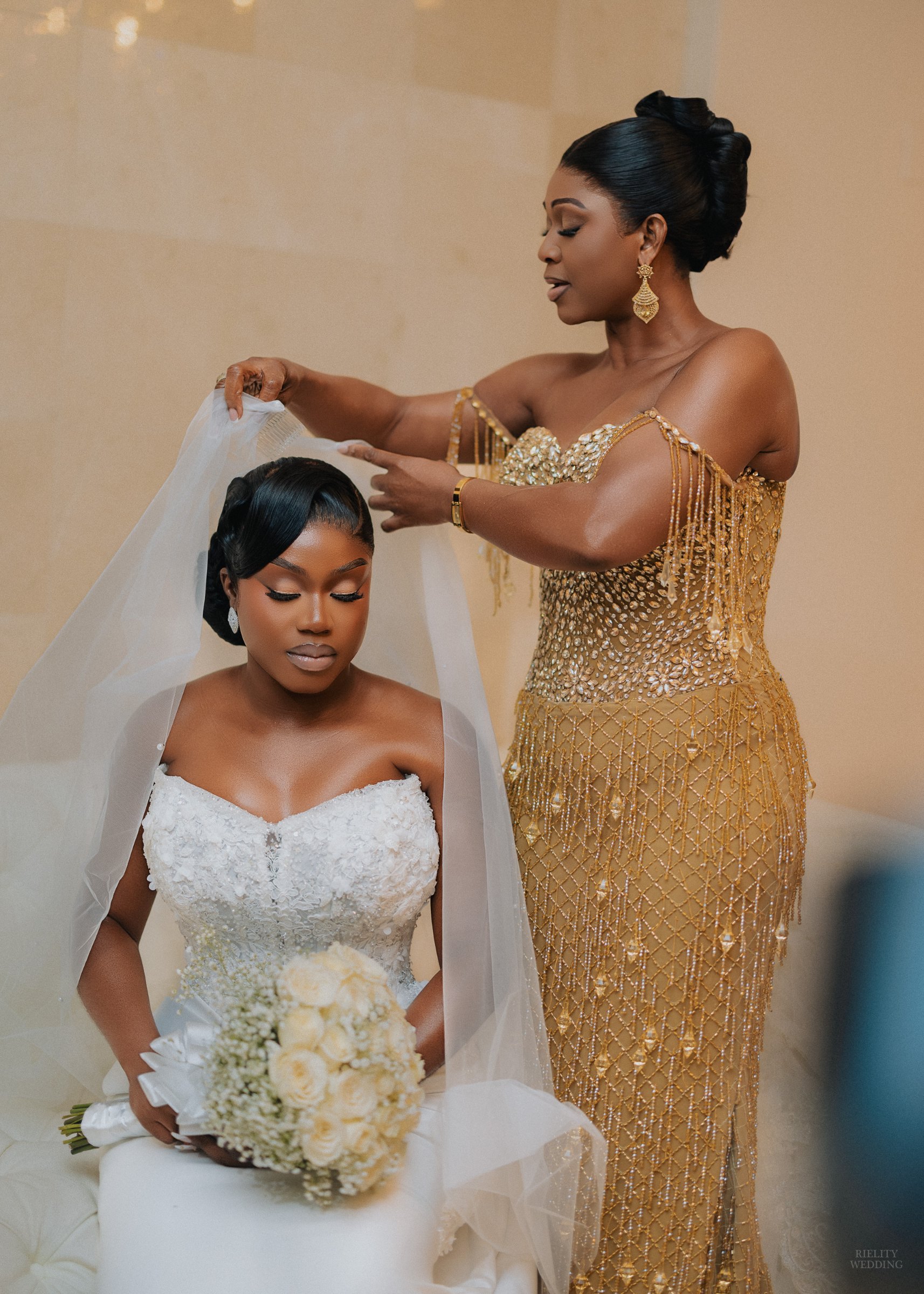 Woman in a gold beaded gown placing a veil over a seated bride holding a white rose bouquet