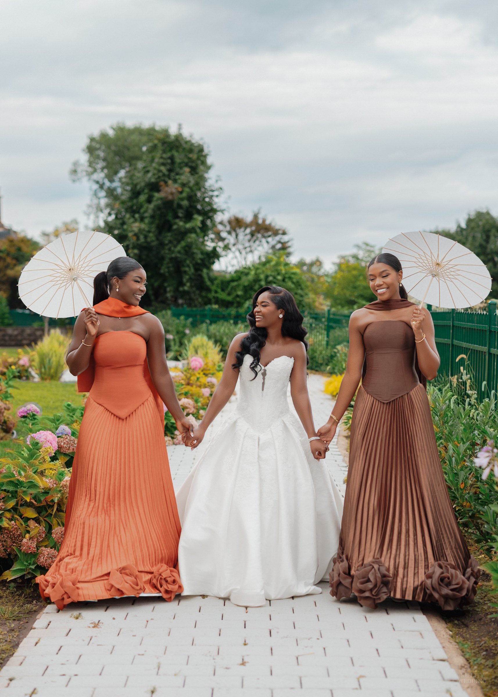 Bride holding hands with two bridesmaids in pleated orange and brown gowns carrying white parasols in a garden