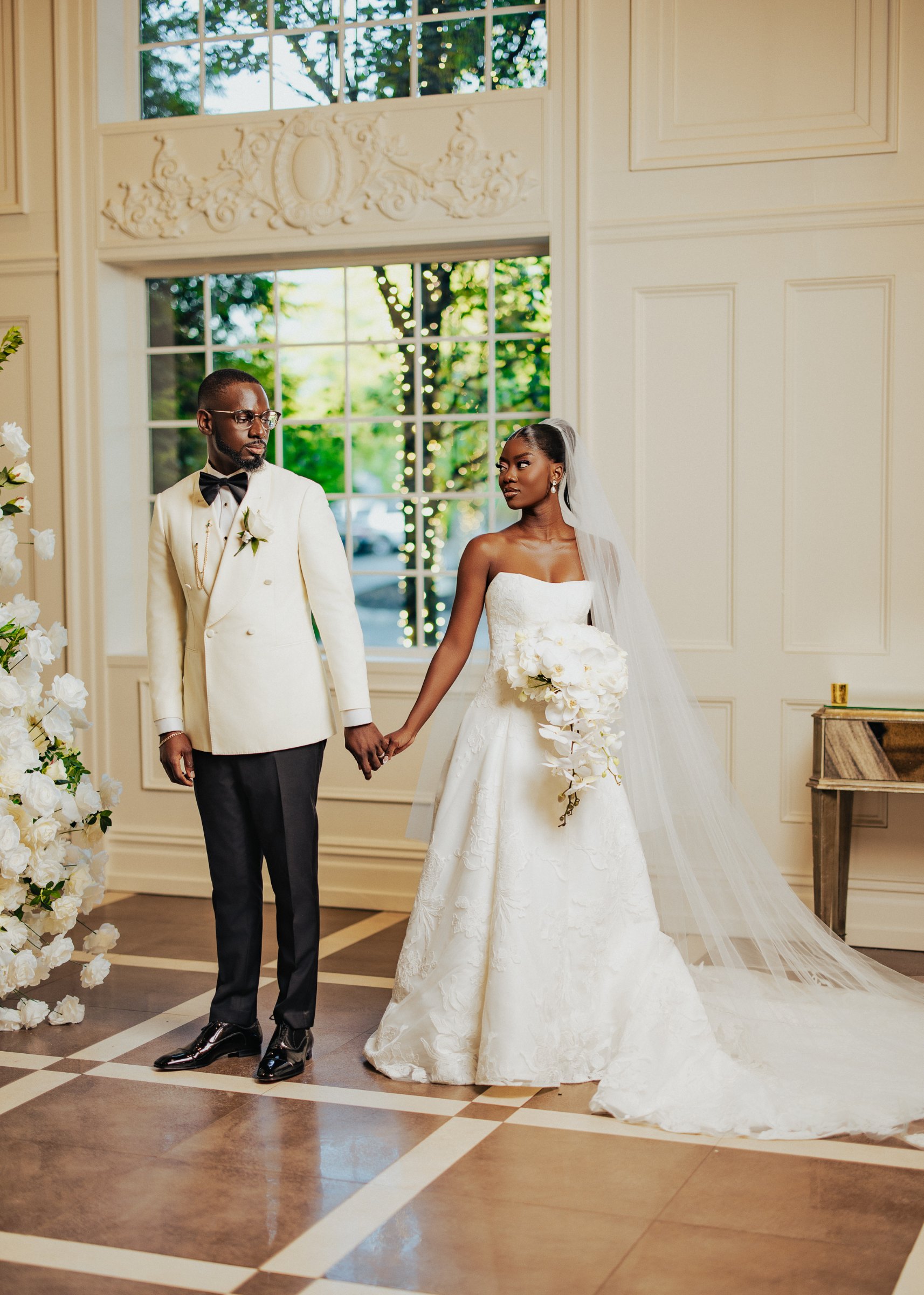 Bride and groom holding hands in an elegant ballroom, groom in an ivory tuxedo jacket and bride with a white orchid bouquet
