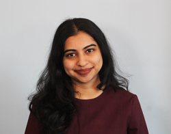 A smiling woman with her hair down wearing a black shirt with white dots on stood in front of a light blue background