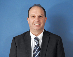 A smiling man in a dark suit with a navy blue tie with light blue stripes stood in front of a dark blue background