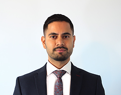 A smiling man with short hair and facial hair with a grey sweater, white shirt and blue tie stood in front of a light blue background.