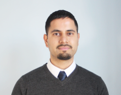 A smiling man with short hair and facial hair with a grey sweater, white shirt and blue tie stood in front of a light blue background.