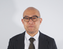 A smiling man with glasses in a black suit, light blue shirt and black tie stood in front of a light blue background