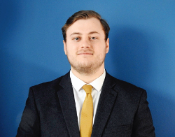 A smiling man with a dark suit on and a yellow tie stood in front of a dark blue background