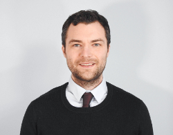 A smiling man with dark hair, a black jumper, burgundy tie and light blue shirt on stood in front of a light blue background