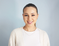 A smiling woman with slicked back hair in a ponytail with a white cardigan and white top on with a gold necklace and earrings stood in front of a light blue background