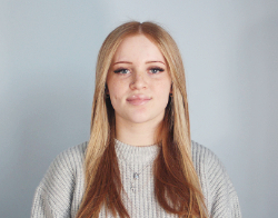 A smiling woman with long hair wearing a grey jumper and a silver necklace stood in front of a light blue background