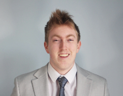 A smiling man with spiked up hair in a grey suit, white shirt and grey tie stood in front of a light blue background