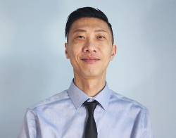 A smiling man with black hair and a blue shirt with a black tie on stood in front of a light blue background